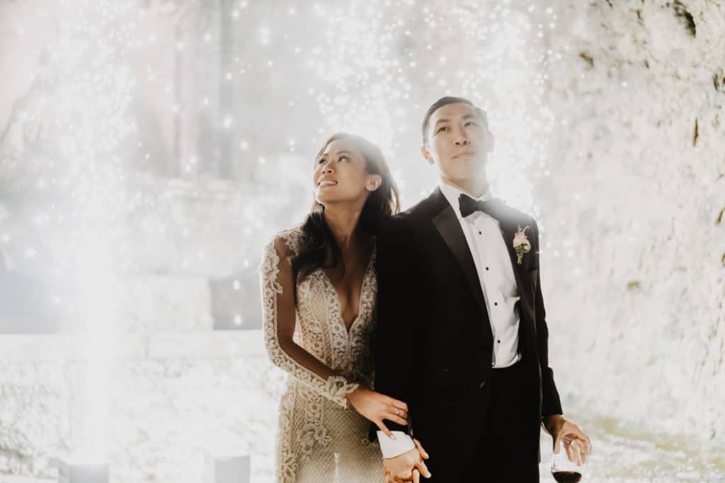 Bride and groom holding each other hands while cold fireworks are shining behind them - Italian Wedding Planners