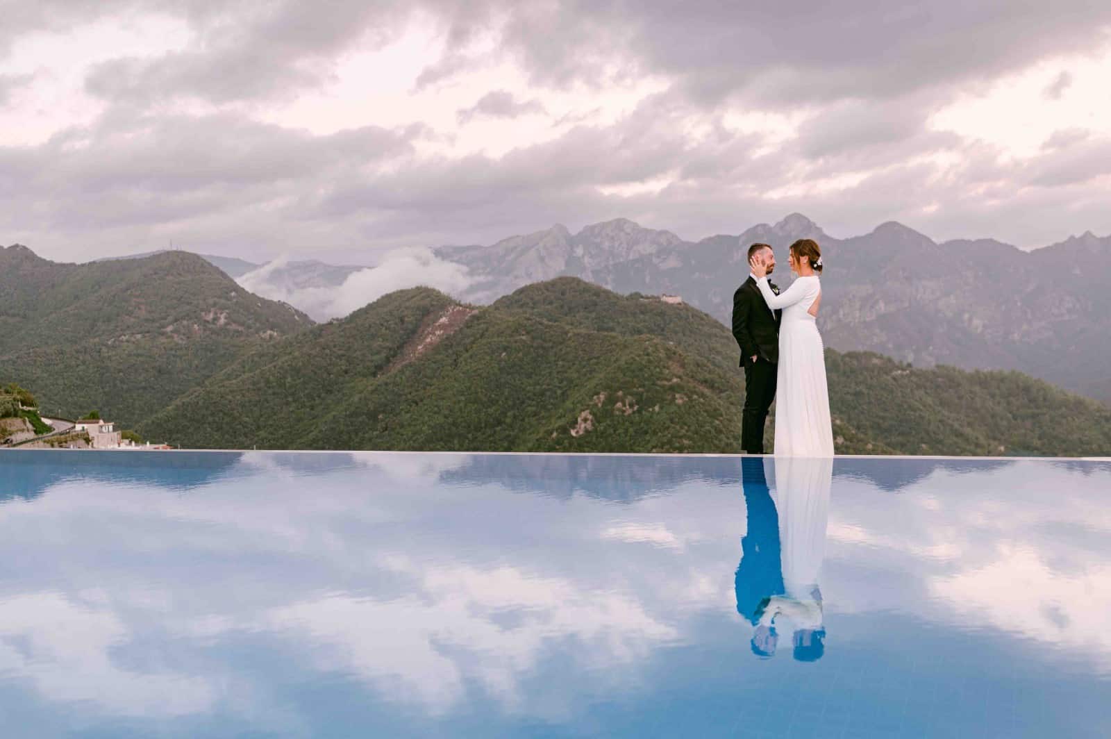 Spouses hug each other near the pool with a view of green hills behind them