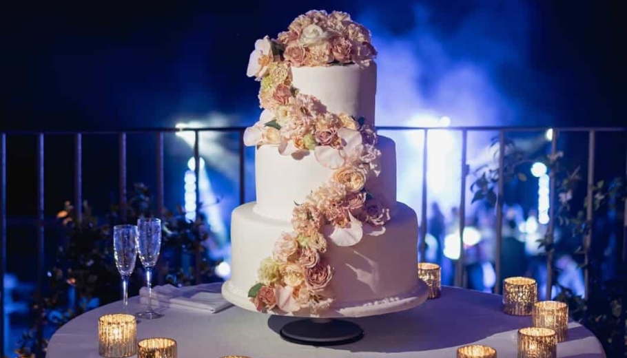 A wedding cake adorned with colorful flowers, displayed on a floral-filled table at a garden party.