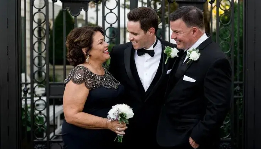 The bride and groom stand with their parents, all smiling happily in a wedding photo, celebrating their union.