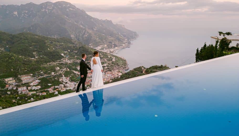 Bride and groom hold hands near an infinity pool overlooking Amalfi Coast scenery.