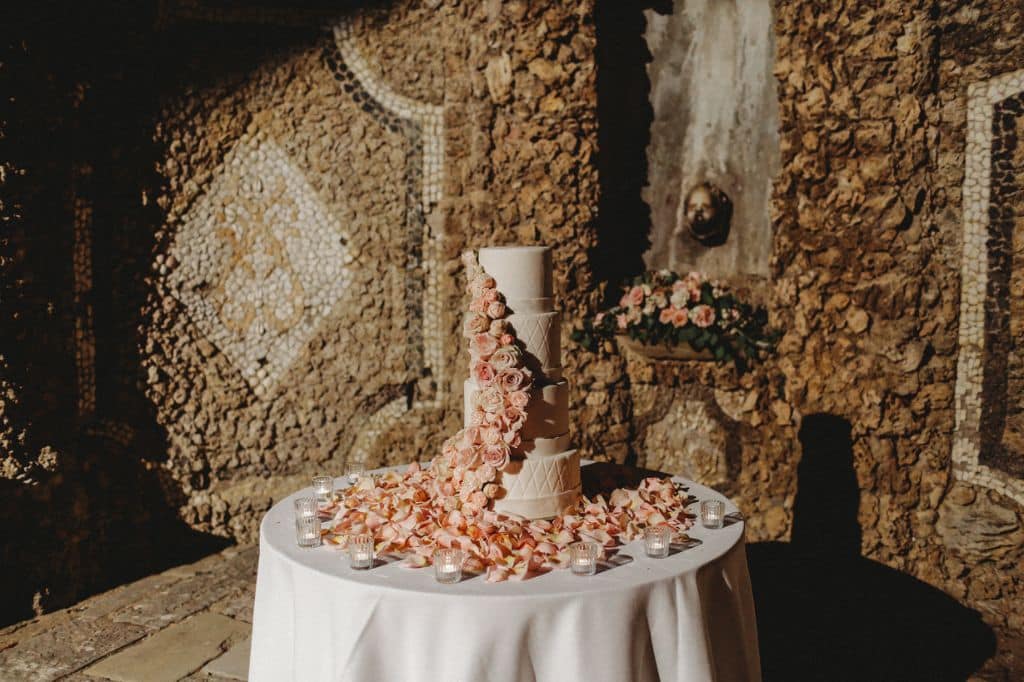 Elegant wedding cake adorned with roses, set against a rustic Italian stone wall for a timeless feel.