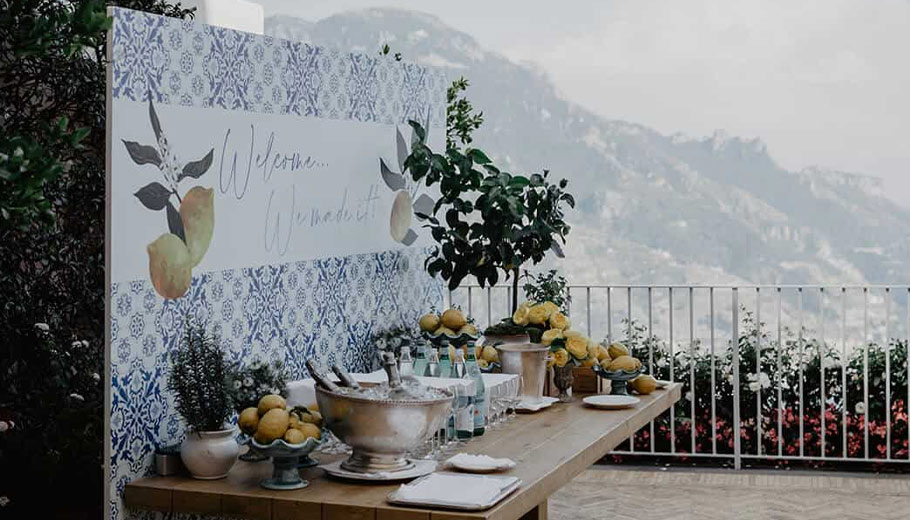 An elegant wedding welcome table set against a backdrop of the Amalfi Coast in Italy, adorned with blue and white decor and fresh lemons, creating a serene and picturesque setting.