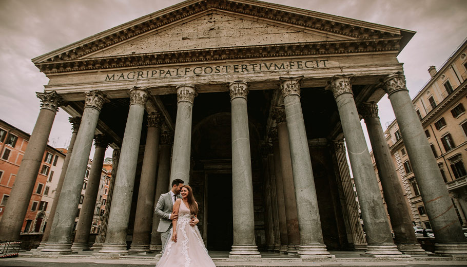 A couple poses in wedding attire in front of the iconic Pantheon in Rome, Italy, under a dramatic cloudy sky.