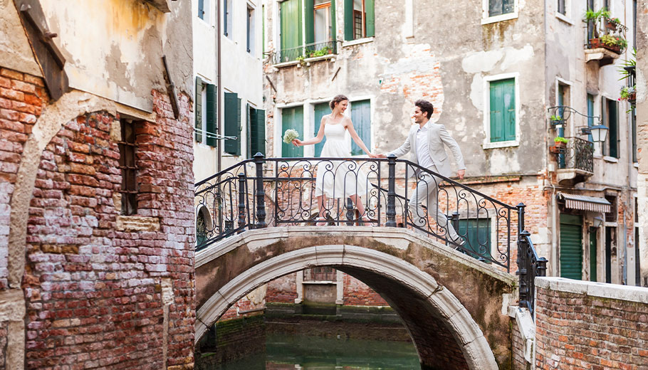 A bride and groom holding hands on a quaint bridge in Venice, surrounded by historic buildings and a canal, capturing a romantic moment in their wedding celebration.