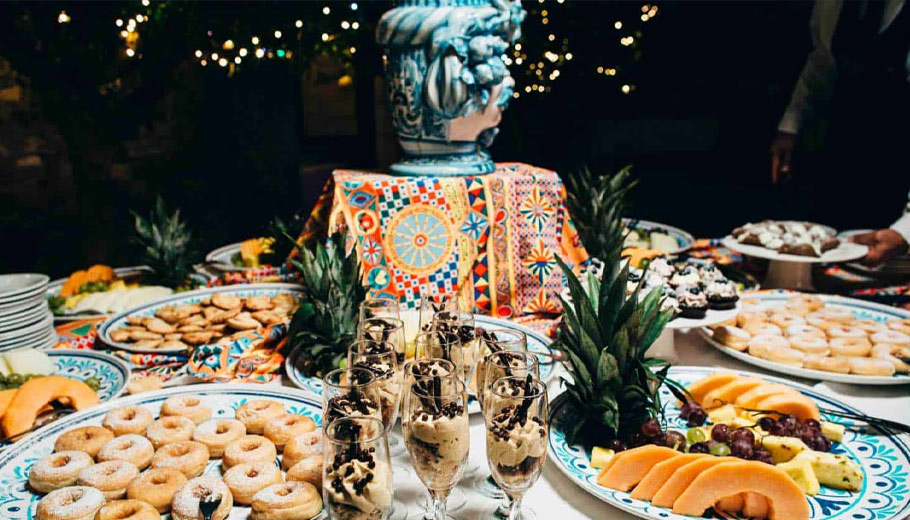 A festive Sicilian wedding buffet with colorful textiles, desserts like donuts and fruit, and sparkling wine glasses.