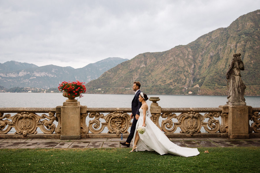 A bride and groom walk along a picturesque lakeside promenade at Lake Como, Italy, with ornate stone balustrades and mountains in the background.